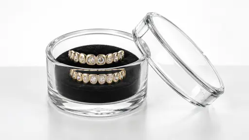 Two plain polished gem set dental grills rest on a dark velvet pad inside an open clear glass storage case with a small lid leaning against it on a white table.