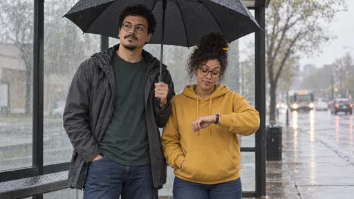 A woman with round tortoiseshell glasses and a curly black bun in a mustard hoodie and a tall man with round glasses and a chin beard in a grey raincoat stand side by side at a bus stop sharing one black umbrella he holds over both of them on a drizzly morning.