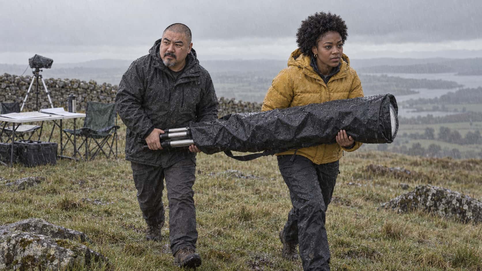A stocky shaven-headed man in a charcoal parka and a young woman in a mustard puffer jacket carry a white telescope wrapped in a waterproof cover between them across a rainy hilltop field with rain streaking diagonally.
