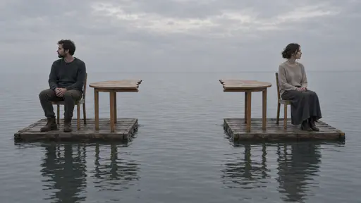 Drifting apart: two adults on chairs aboard separate small rafts drift apart across gray water, a table split in halves facing each other over the gap.