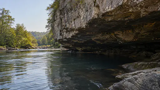 The river runs close under an undercut bank beneath a broad rock overhang in soft even light, the water dark in the overhang shadow.