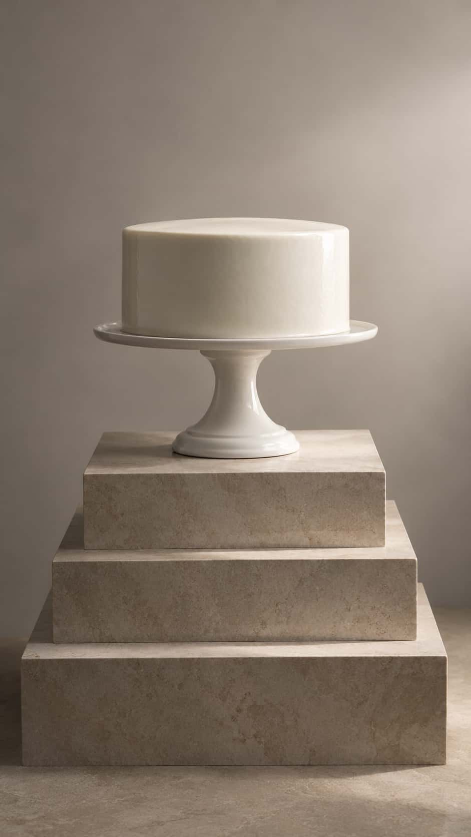A smooth white glazed round cake sits on a tall footed white porcelain cake stand on a stepped honed stone pedestal against a pale warm grey backdrop in a studio photograph.