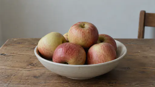 A cluster of whole plain apples balances together in one shallow plain bowl on a plain table in even daylight.