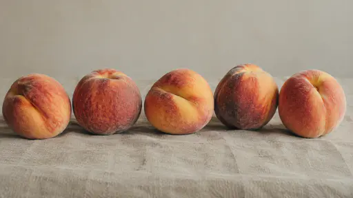 Whole peaches with contrasting intact skins rest in a loose row on a plain linen cloth in soft warm light.