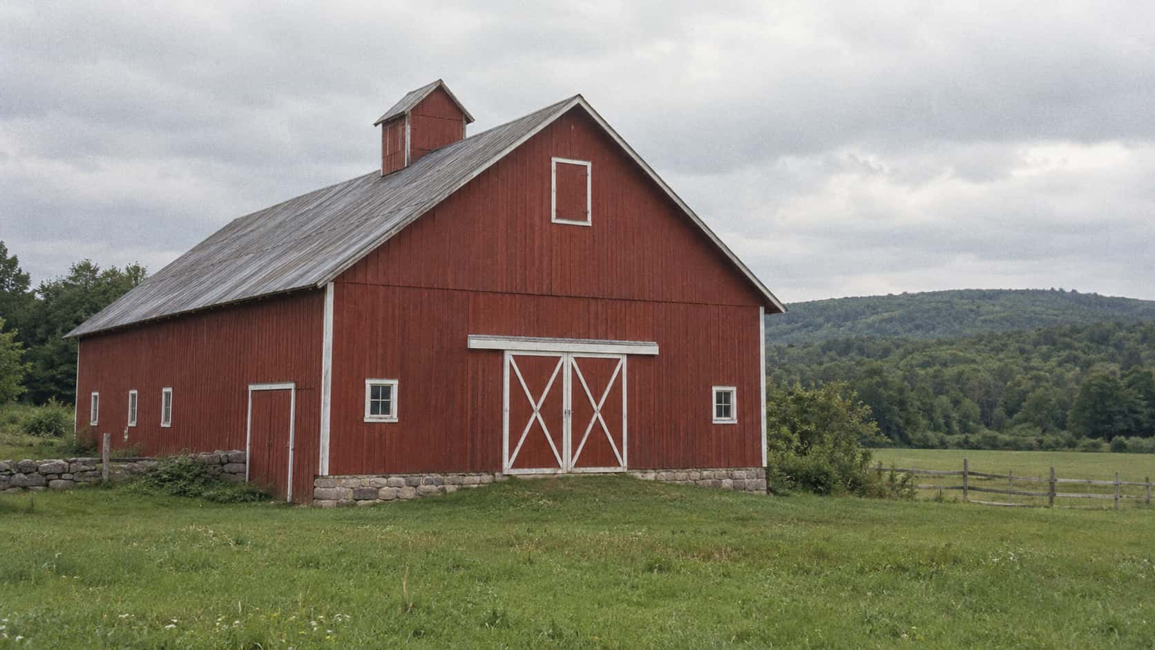 A whole red barn stands beside an open green field with its roofline complete against the sky in even daylight.