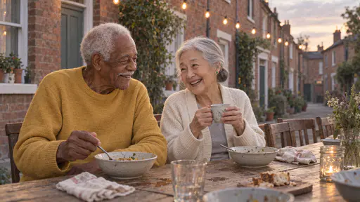 A tall elderly man with white hair and a white moustache and a very small elderly woman with a silver bun share a quiet laugh over half-eaten bowls at a long street table under a string of plain warm bulbs in soft dusk light.