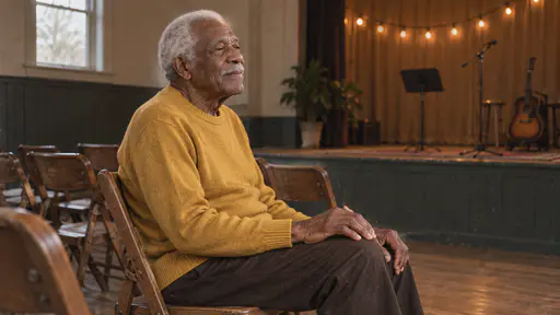 A tall elderly man with close-cropped white hair and a white moustache sits in the front row on a folding chair with his eyes half closed and his hand tapping his knee in time under warm stage light and string bulbs.
