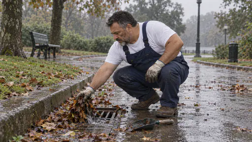 A stocky man with a round face and short grey beard in dark blue overalls and work gloves crouches beside a flooded drain grate on a park path scooping a mat of wet leaves away while rainwater clears into the drain in grey overcast light.