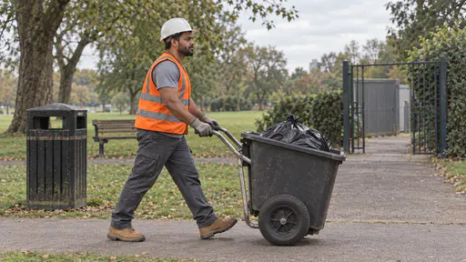 A broad strong man with brown skin and a short black beard in a white hard hat and orange high-visibility vest wheels a wide two-wheeled collection cart holding a full black bin bag away from a litter bin toward a park service gate in bright afternoon light.