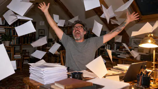 A man in his forties with grey stubble and glasses in a grey t-shirt throws his arms up with a whoop in an attic study as loose manuscript pages fly through the air.